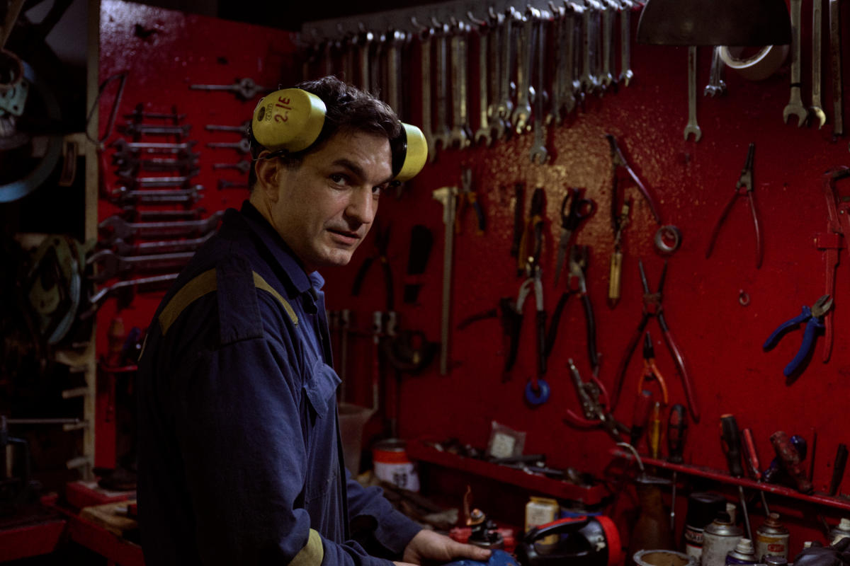Ein Seemann in dunkelblauem Overall steht vor der Werkbank. Er trägt hochgeklappte gelbe Ohrschützer. An der Wand hängen viele Werkzeuge.
